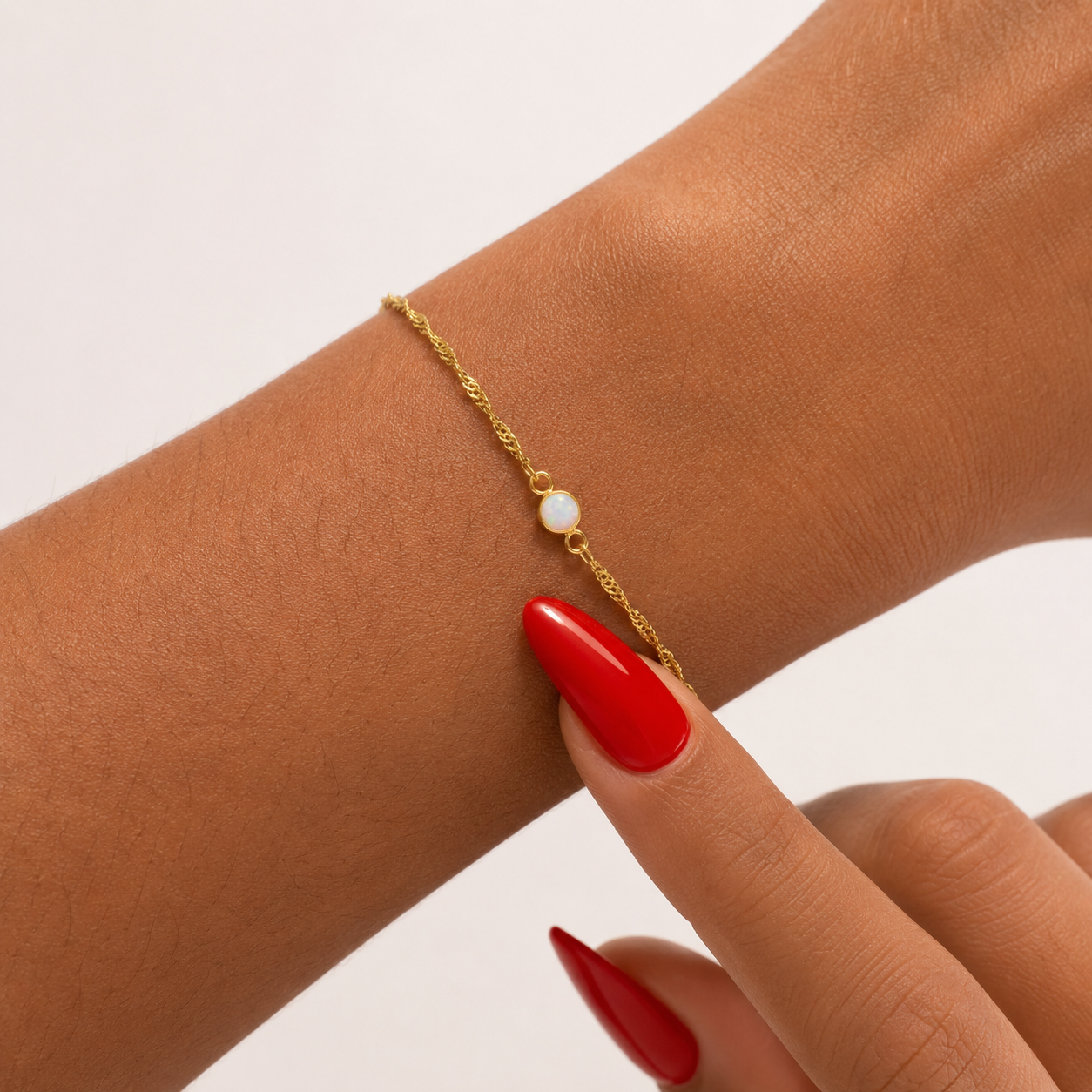 Gold bracelet with a opal charm on a wrist with red nail polish against a light background