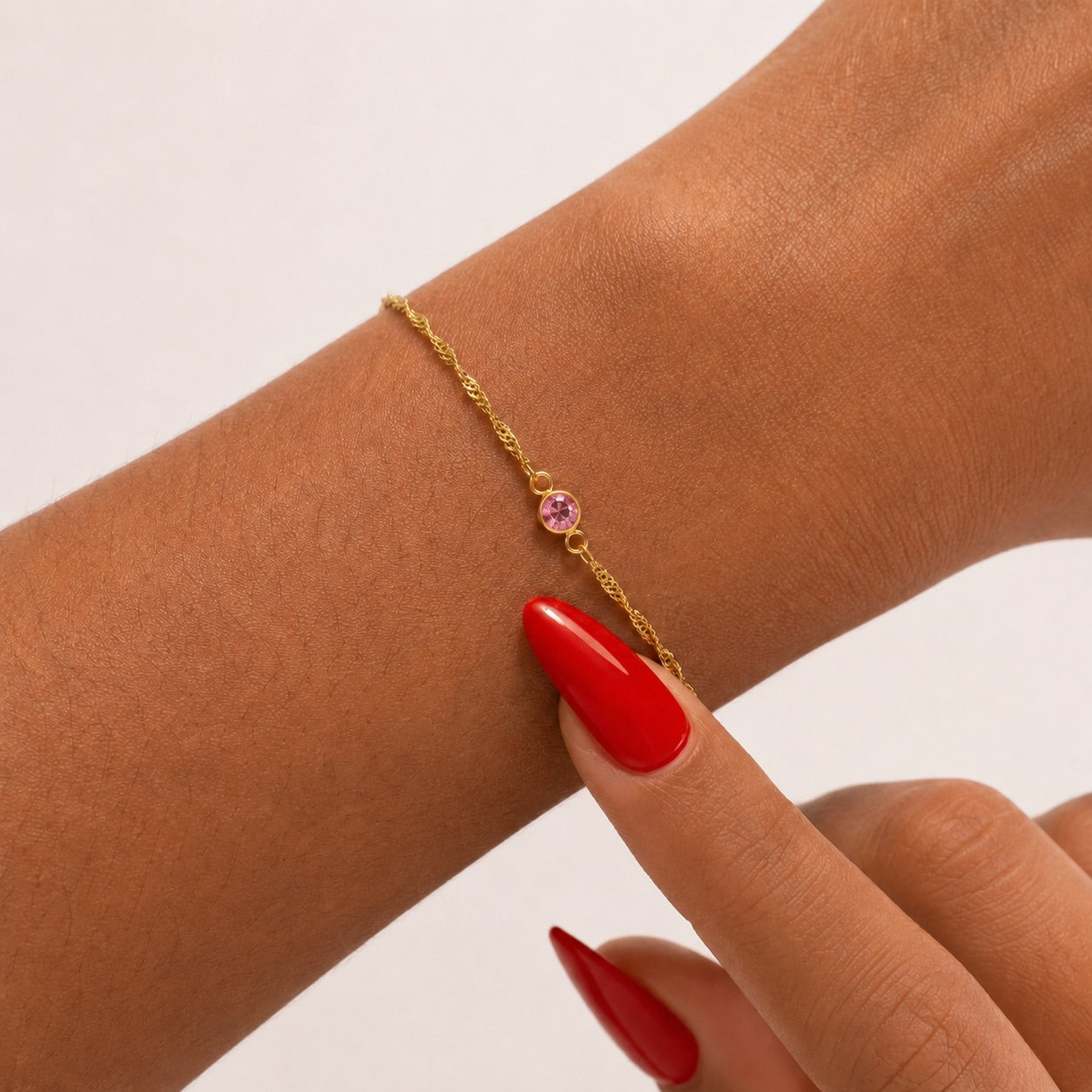 Gold bracelet with a pink gemstone on a wrist with red nail polish against a neutral background
