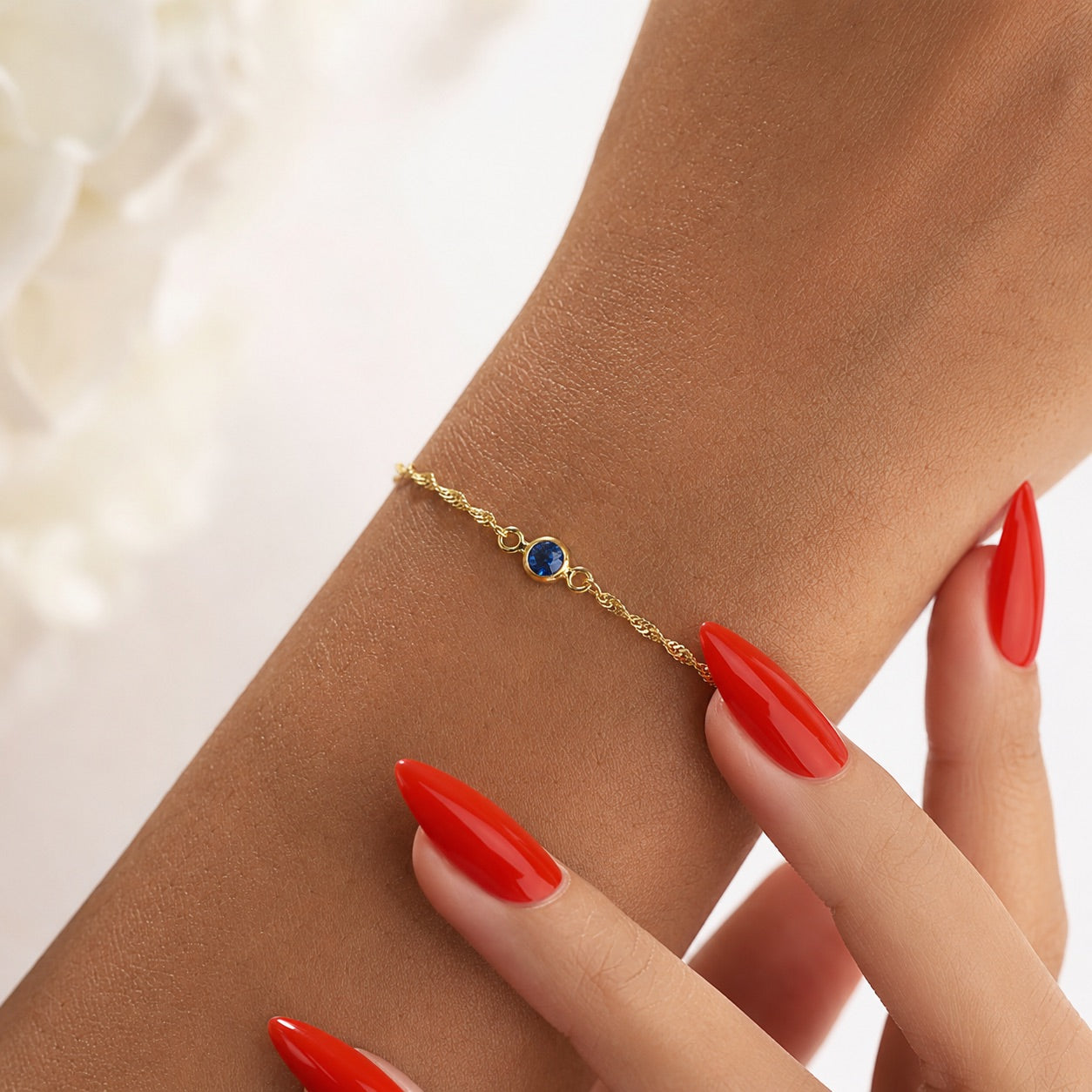 Gold charm bracelet with a September blue gemstone on a wrist with red nail polish.